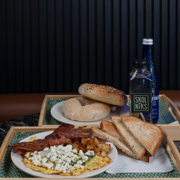 Breakfast tray with omelet, bacon, potatoes, toast, bagels, and bottled water on a dark table.