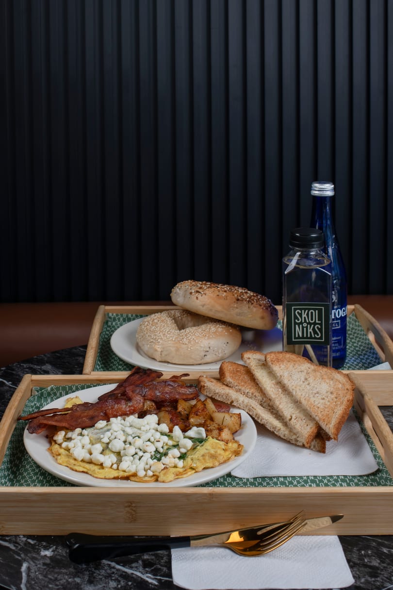 Breakfast tray with omelet, bacon, potatoes, toast, bagels, and bottled water on a dark table.