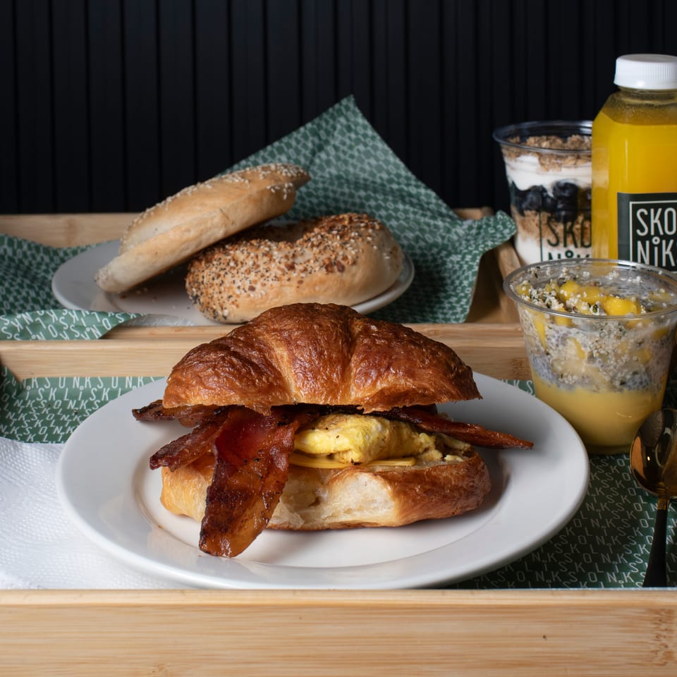 Breakfast tray with a bacon, egg, and cheese croissant, bagels, yogurt, chia pudding, and orange juice.
