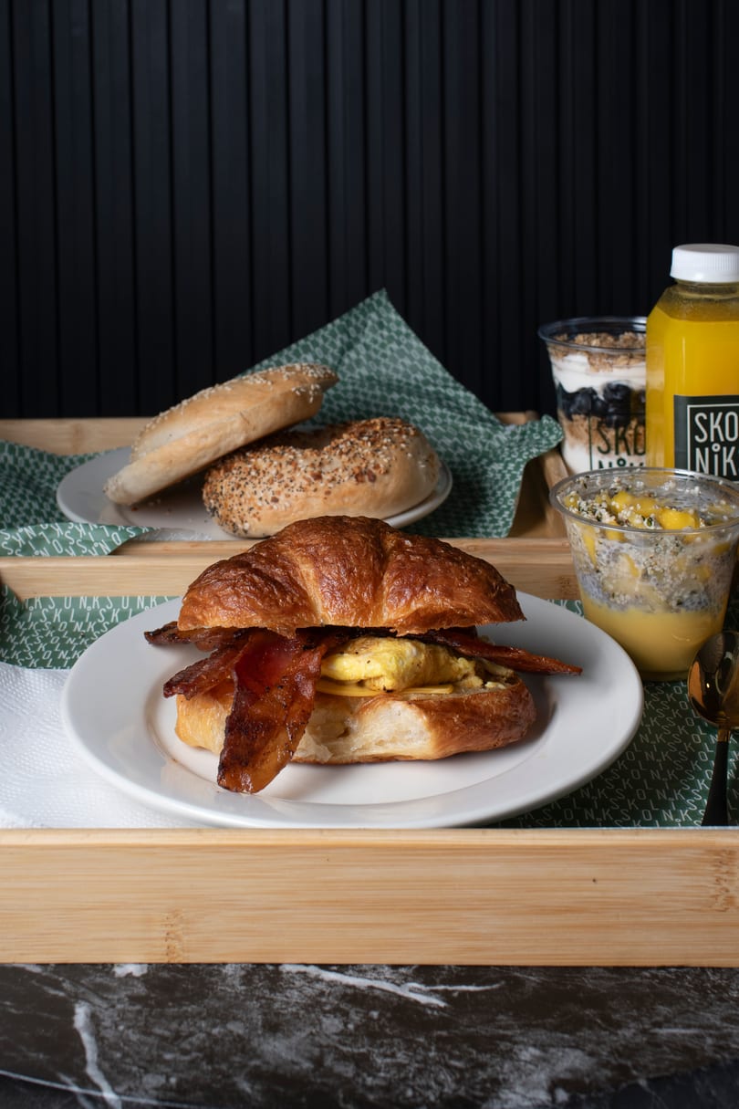 Breakfast tray with a bacon, egg, and cheese croissant, bagels, yogurt, chia pudding, and orange juice.