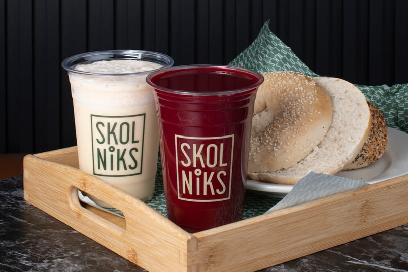 Two Skol Niks branded drinks, one white and one red, with sliced bagels on a wooden tray.