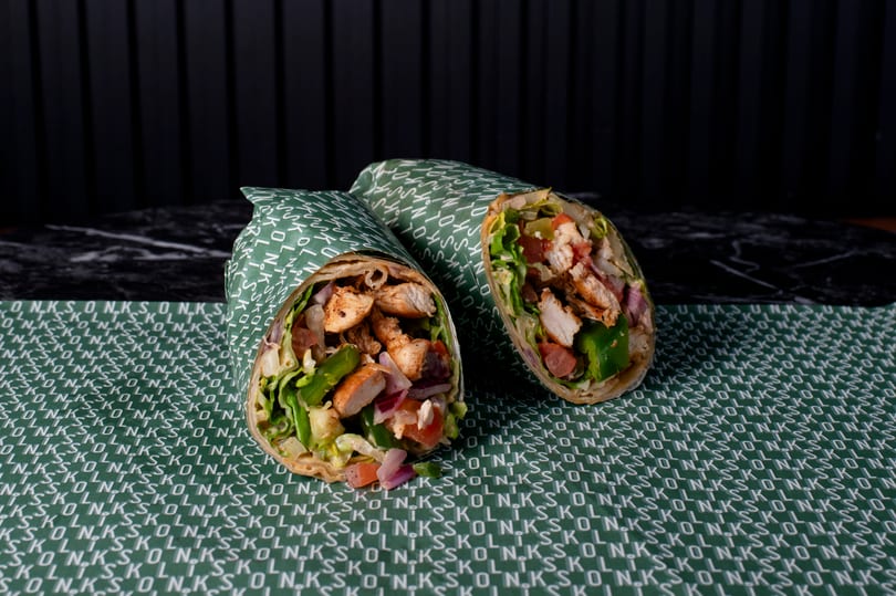 Two chicken wraps, filled with lettuce, tomatoes, and onions, on green branded paper.