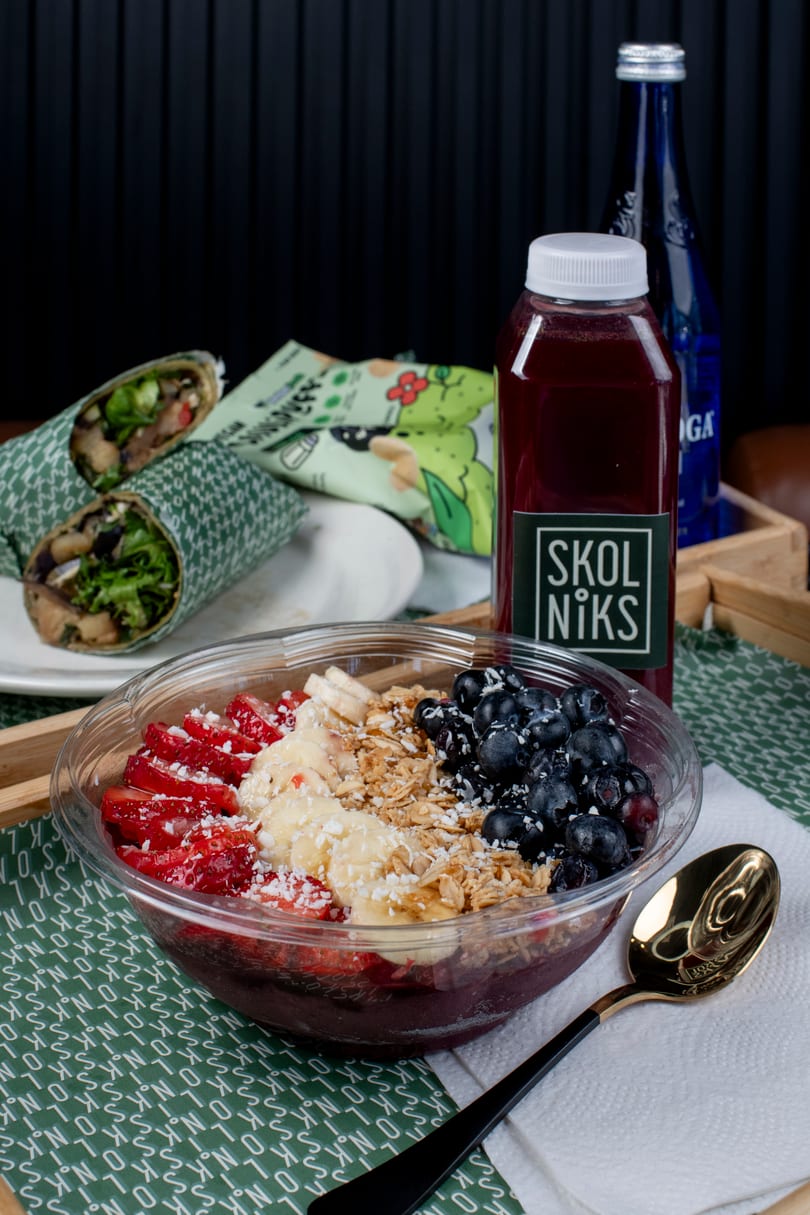 A Skolniks acai bowl with strawberries, bananas, blueberries, and granola, next to wraps and drinks.